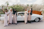 the bridesmaids around the Cadillac holding flowers in the air as the bride looks out of the car window with her bouquet